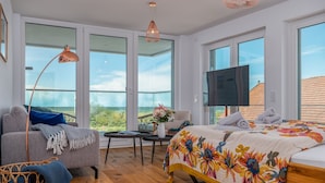 A modern bedroom with a large bed, a sofa, a TV, and a balcony with a view.