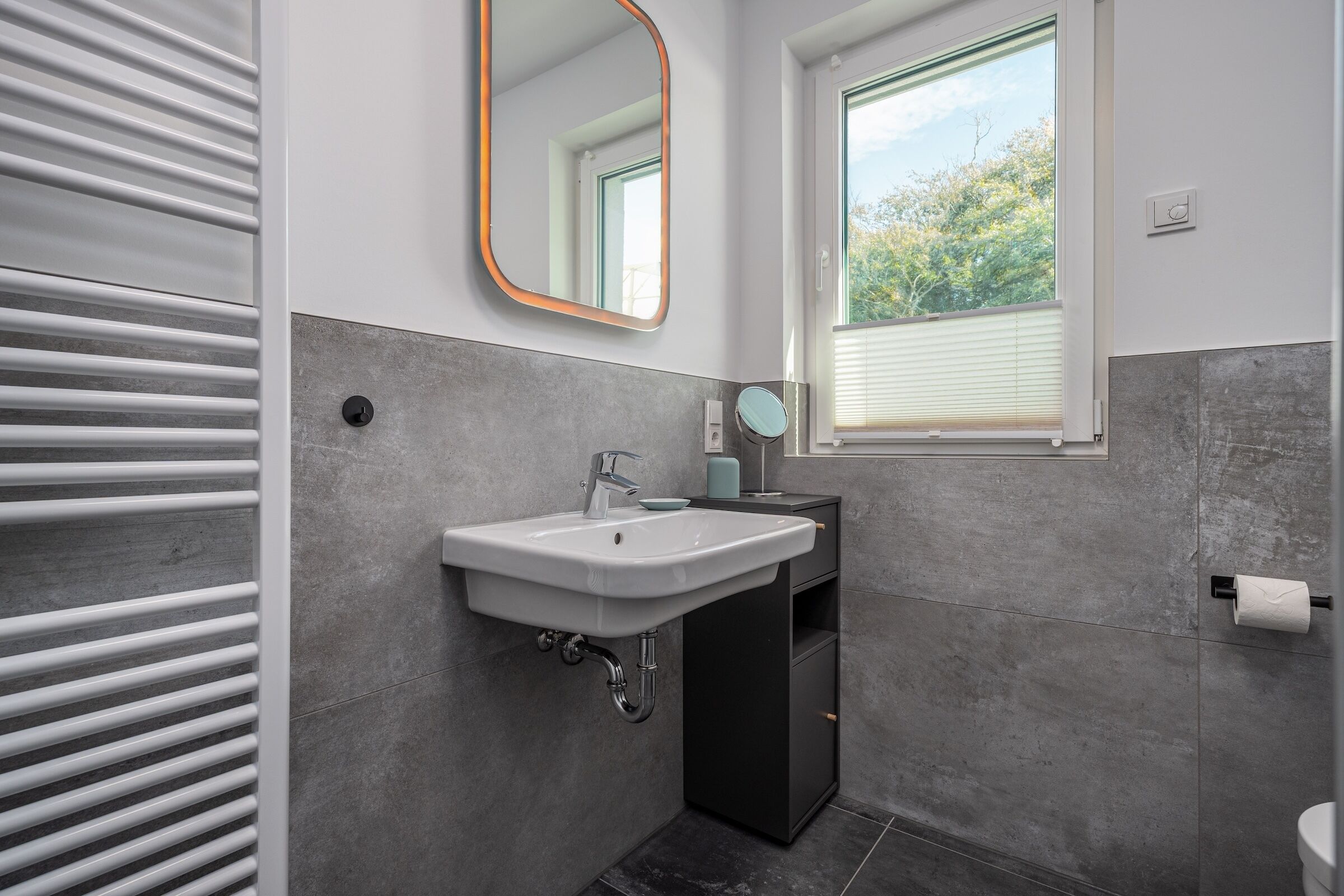 A modern bathroom with a white sink, dark cabinet, and a window with blinds.