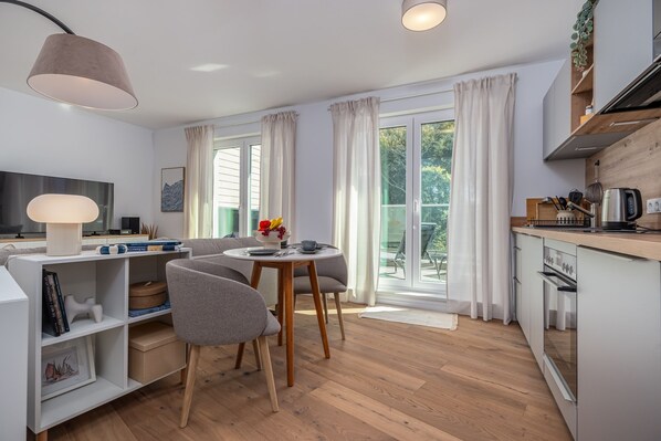 A modern kitchen with a dining area, wooden flooring, and a large window with curtains.