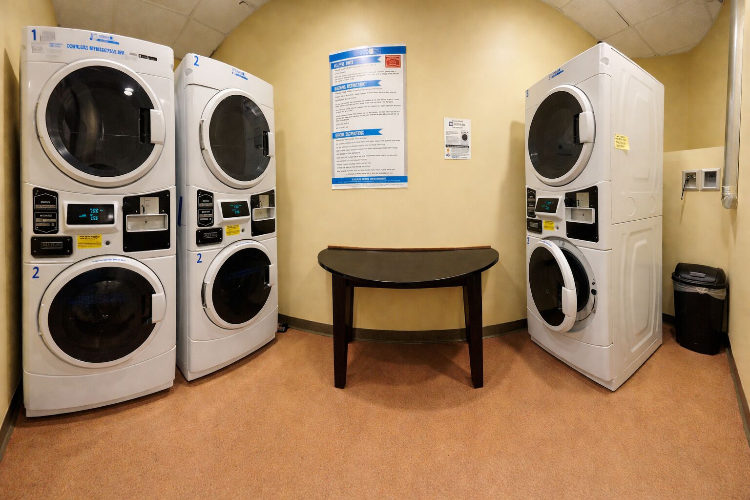 Een wasserij met drie gestapelde wasmachines en drogers, een klein tafeltje en informatieposters aan de muur.