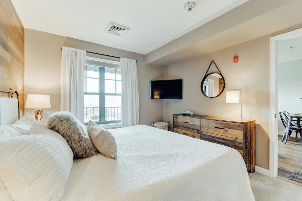 A bedroom with a large bed, a wooden dresser, a flat-screen TV, and a window with curtains.