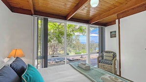 A room with a bed, a chair, a lamp, and large windows offering a view of the outdoors.