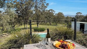 A picnic setup with a bottle of wine, glasses, and a fruit platter on a wooden table, overlooking a pool and lush greenery.