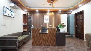 A reception area with a wooden reception desk, a sofa, a ceiling fan, and a potted plant. - Hotel Raj Mahal (Gurugram)