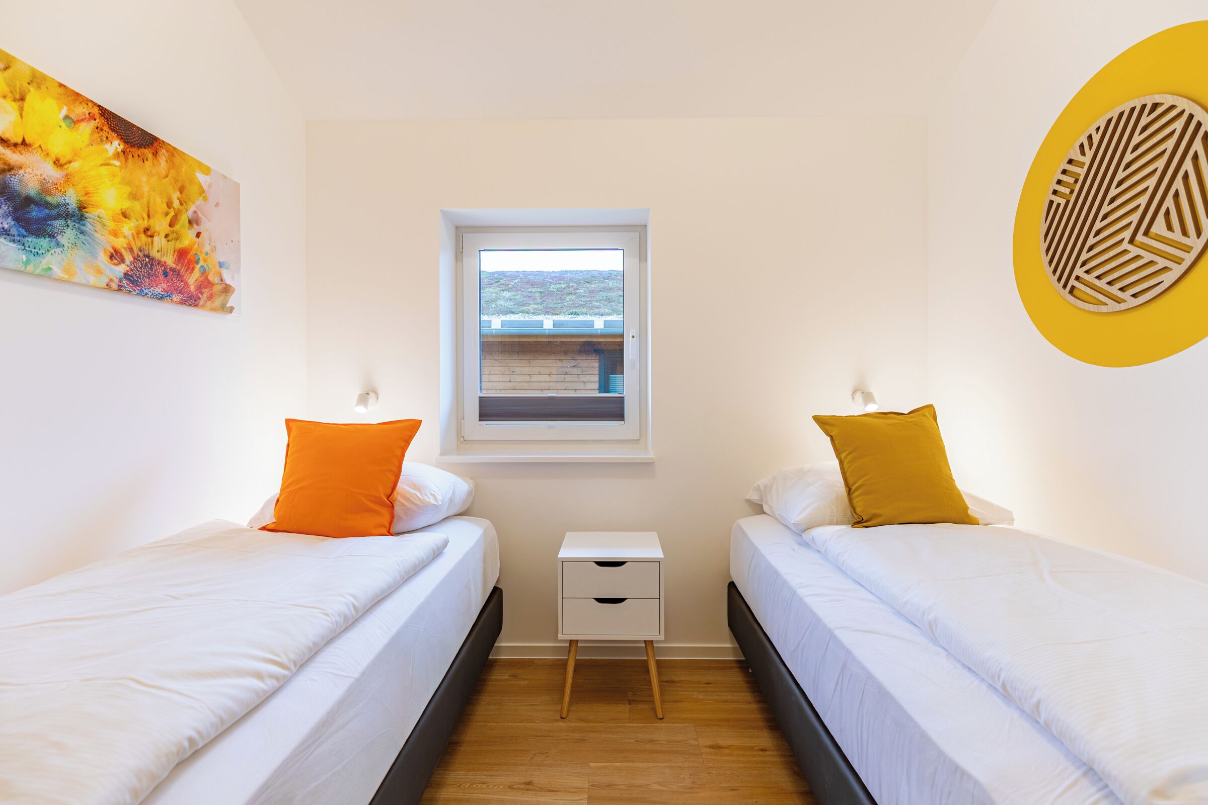 A bedroom with two single beds, a small bedside table, a window with a view, and a colorful abstract painting on the wall.