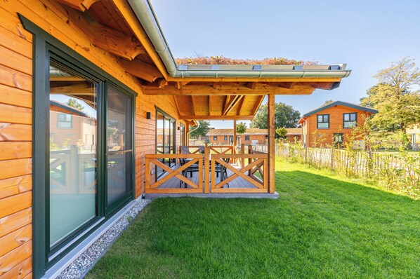 A wooden cabin with a covered patio area, a grassy yard, and a neighboring building.