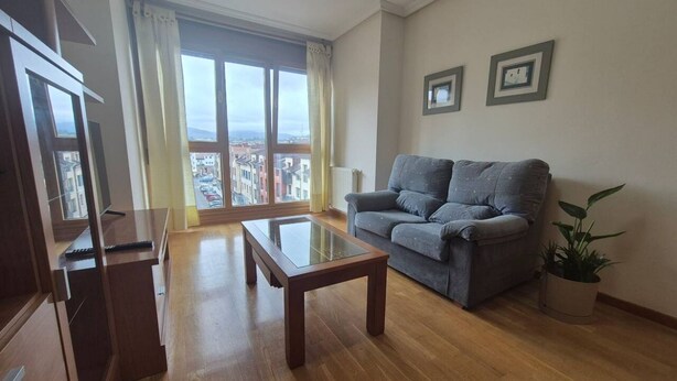 Un salon avec un canapé, une table basse en verre et une vue sur les immeubles par une grande fenêtre.