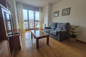A living room with a sofa, a glass-top coffee table, and a view of buildings through a large window.