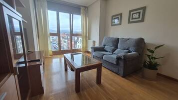 A living room with a sofa, a glass-top coffee table, and a view of buildings through a large window.