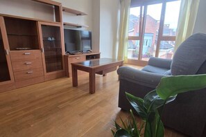 A living room with a sofa, a television on a stand, a wooden cabinet, and a window with a view of buildings.