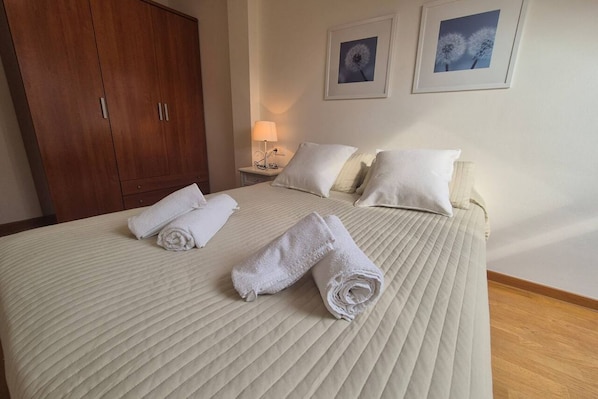 A neatly made bed with rolled towels, a wooden wardrobe, a bedside table with a lamp, and two framed pictures on the wall.