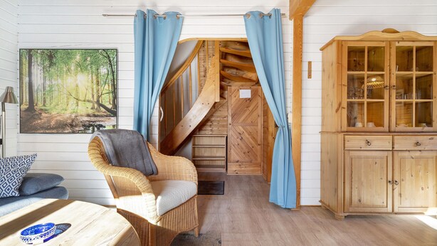 Una habitación con escalera de madera, una silla de mimbre, un armario y un cuadro de un bosque.