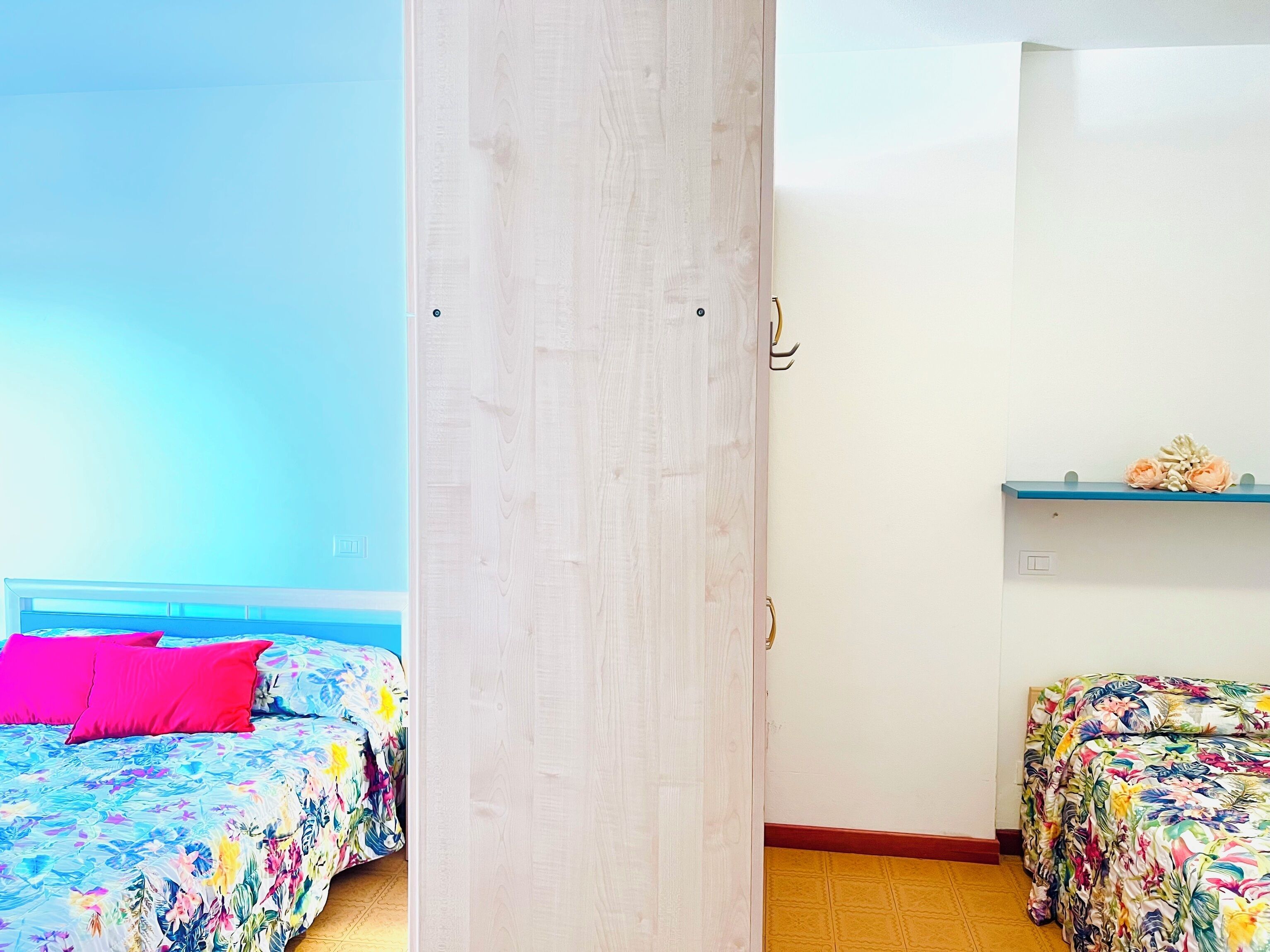 A room with two beds, a wooden door, a shelf with flowers, and a wall switch.