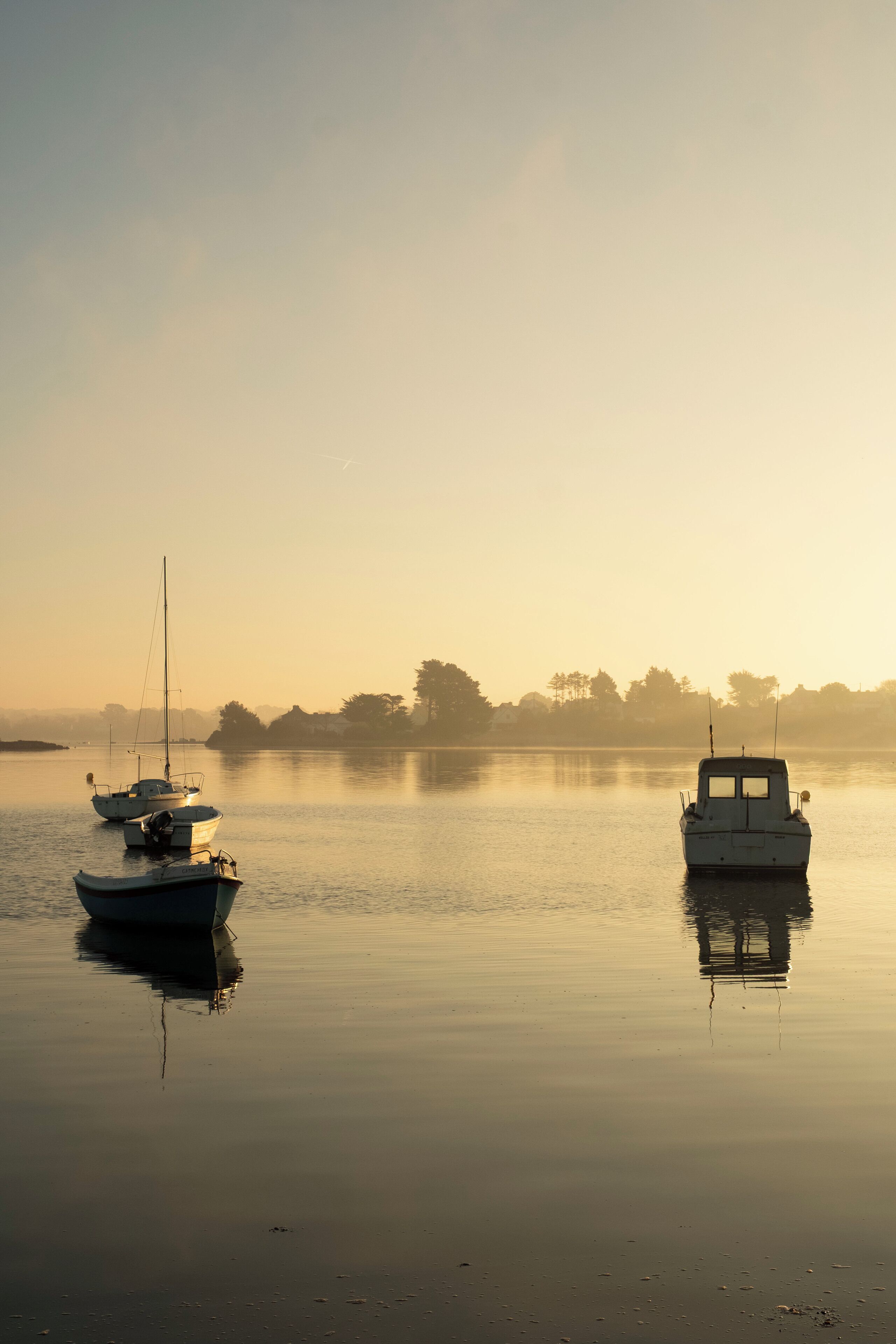 A calm lake with two boats anchored, surrounded by trees and a misty horizon.