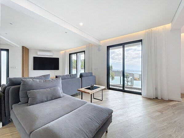 A modern living room with a grey sofa, a flat-screen TV, and a balcony with a view of the sea.