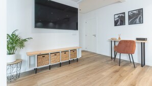 A modern living room with a large flat-screen TV mounted on the wall, a wooden bench with storage baskets, a dining table with a chair, and two framed pictures on the wall. - Breeze House 2 by Atlantic Holiday (Caniço)
