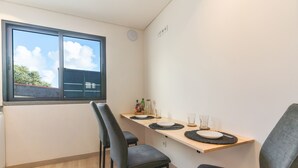 A small dining area with a table set for two, two chairs, a window with a view of the sky and trees, and a wall-mounted electrical outlet. - Breeze House 2 by Atlantic Holiday (Caniço)