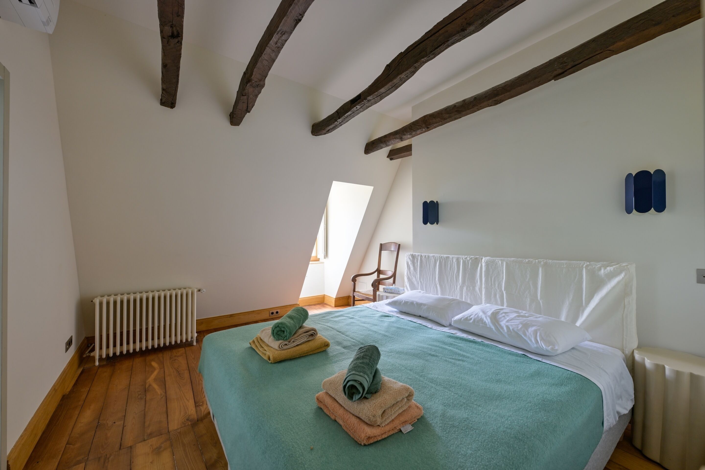 Une chambre avec un plancher en bois, un lit recouvert d’une couverture verte et une tête de lit blanche. Il y a un radiateur au mur et une fenêtre donnant sur l’extérieur.