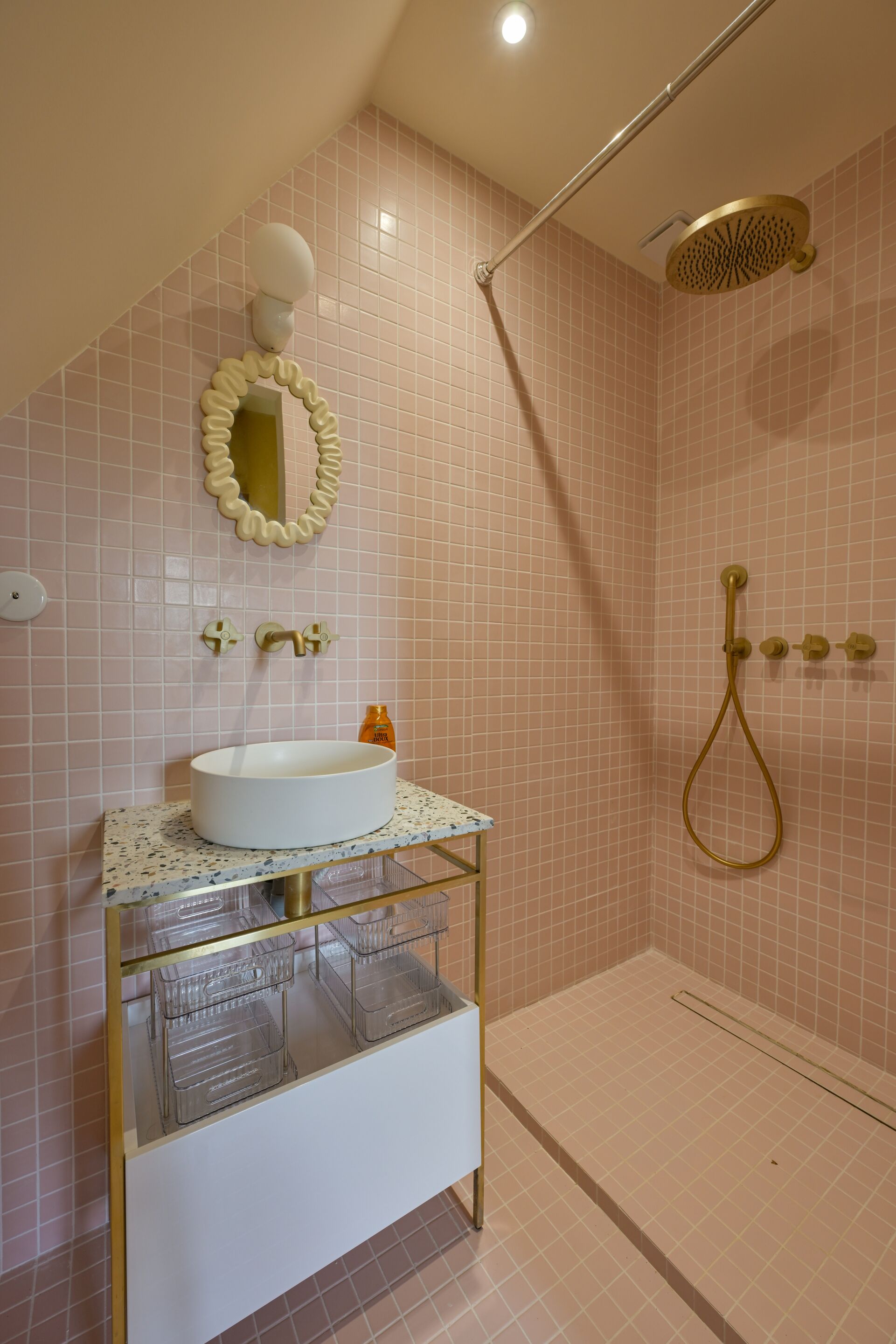 Une salle de bain avec un lavabo blanc, un meuble vitré, un miroir avec un cadre décoratif, une douche avec un pommeau de douche doré et des murs recouverts de carreaux roses.