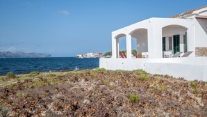 A coastal building with white walls and green shutters, a patio with seating, and a view of the sea and distant buildings. - Villa 'Punta Marina' with sea views, private terrace and Wi-Fi (Illes Balears)