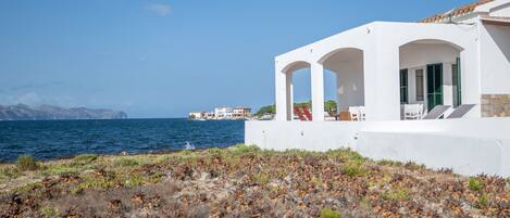 A coastal building with white walls and green shutters, a patio with seating, and a view of the sea and distant buildings.