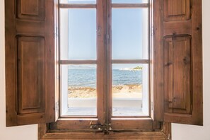 A wooden-framed window with open shutters, offering a view of a beach and distant land.