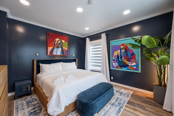 A modern bedroom with a large bed, two paintings, a potted plant, and a nightstand.