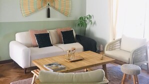 A living room with a sofa, a coffee table, a wicker chair, a fan, and a window with curtains. - Gîte / furnished 3-bedroom house with veranda and garden - available 13/09 → 15/10 (Lagor)