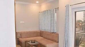 A modern living room with a beige sectional sofa, a small wooden side table, and a large window with light curtains. - Hotel The Lake Castle Ranchi Jharkhand (Ranchi)