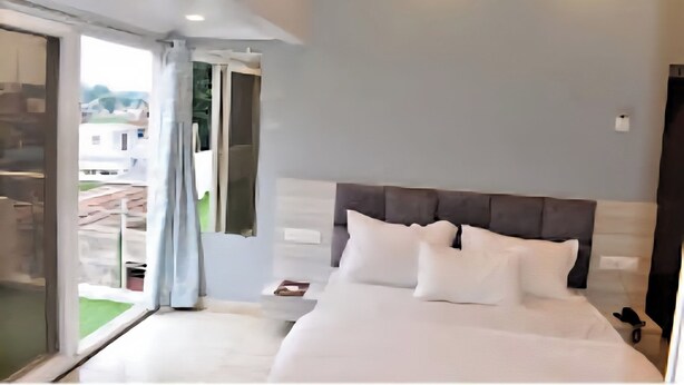 A modern bedroom with a large bed, a glass door leading to a balcony, and a view of buildings outside. - Hotel The Lake Castle Ranchi Jharkhand (Ranchi)