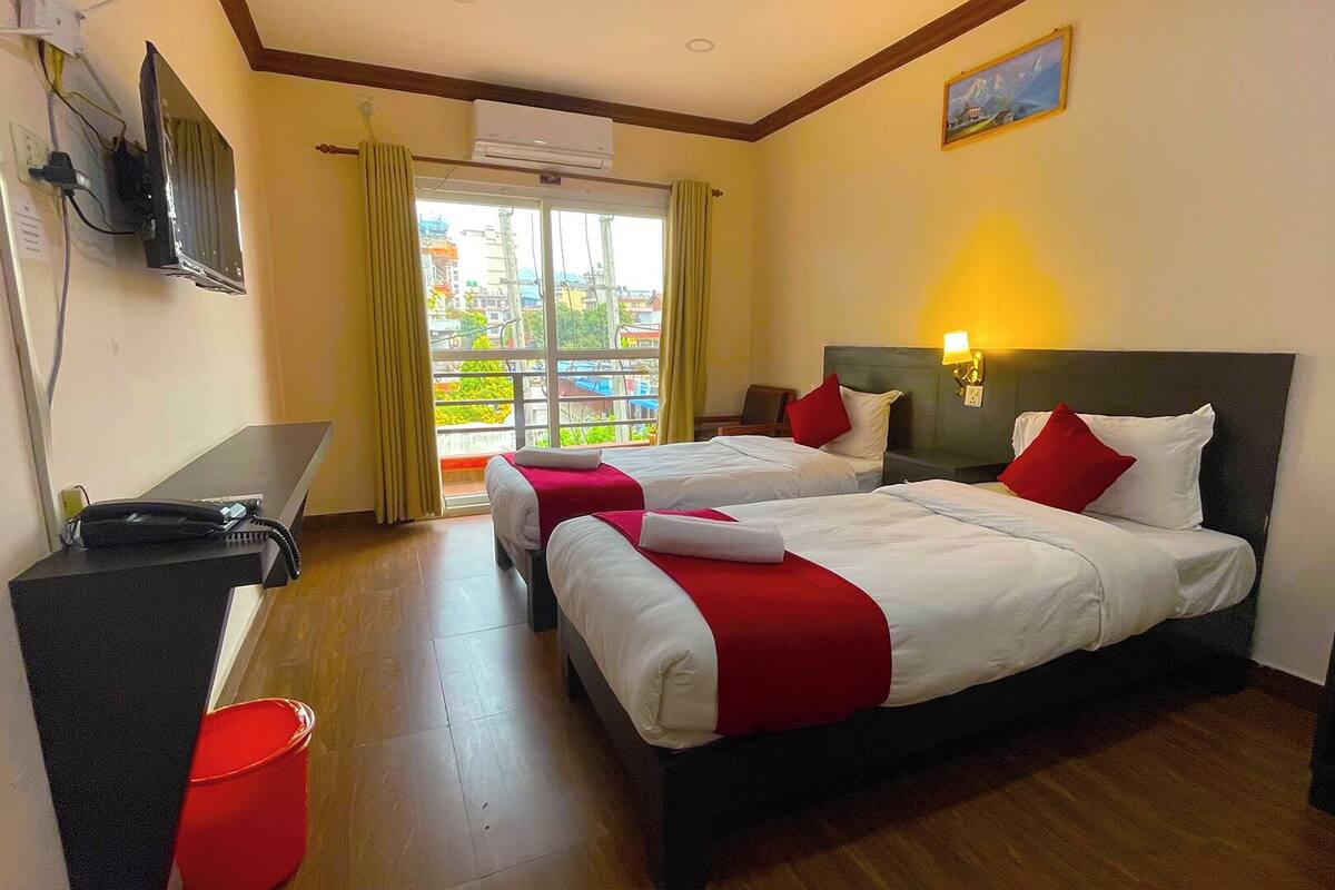 A hotel room with two beds, a desk, a TV, and a window with a view of buildings and greenery.