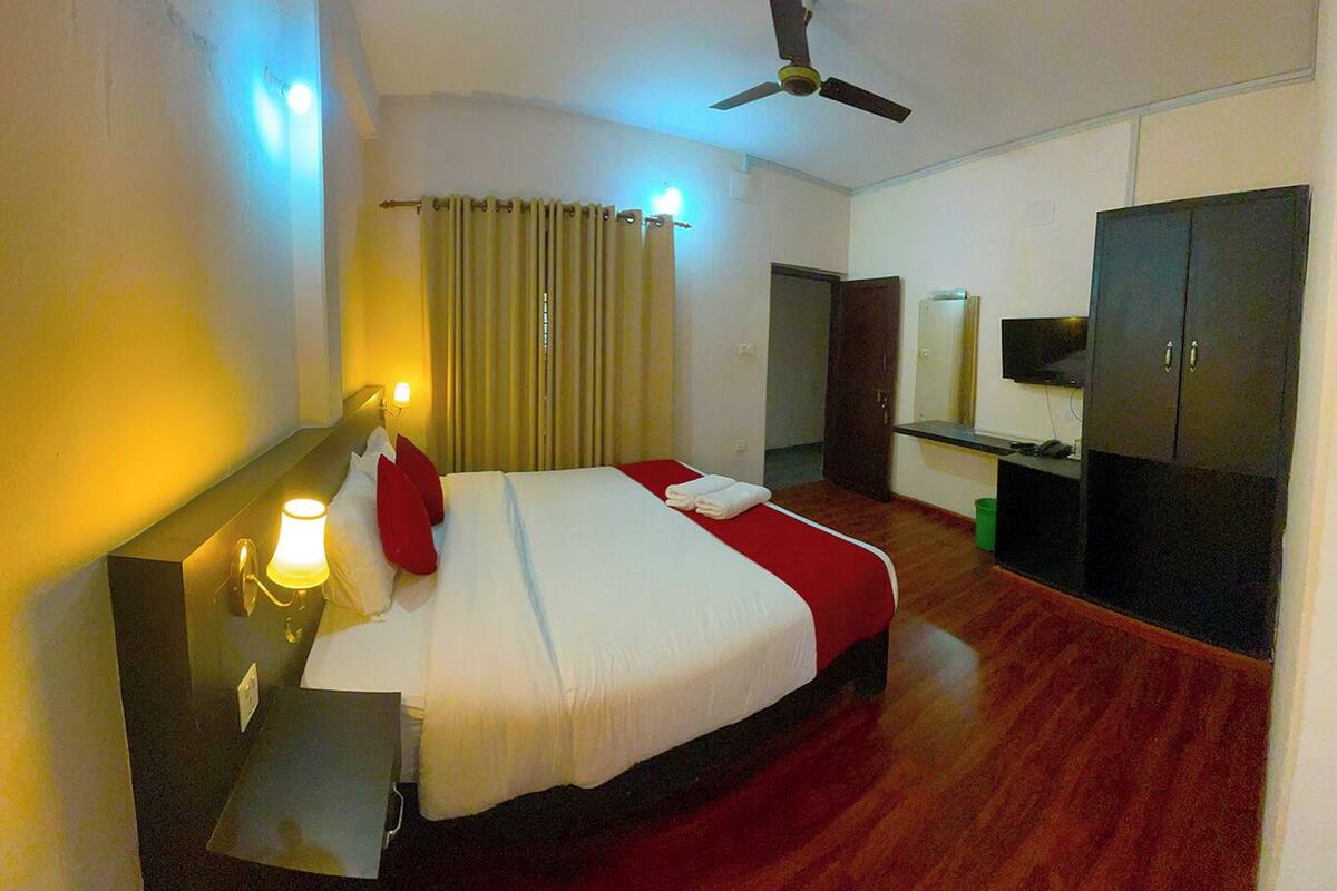 A hotel room with a bed, bedside table, lamp, curtains, ceiling fan, wardrobe, and a television.