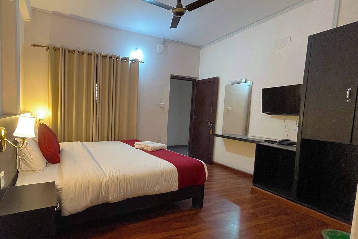 A hotel room with a bed, a desk with a lamp, a wardrobe, a television, and a ceiling fan.