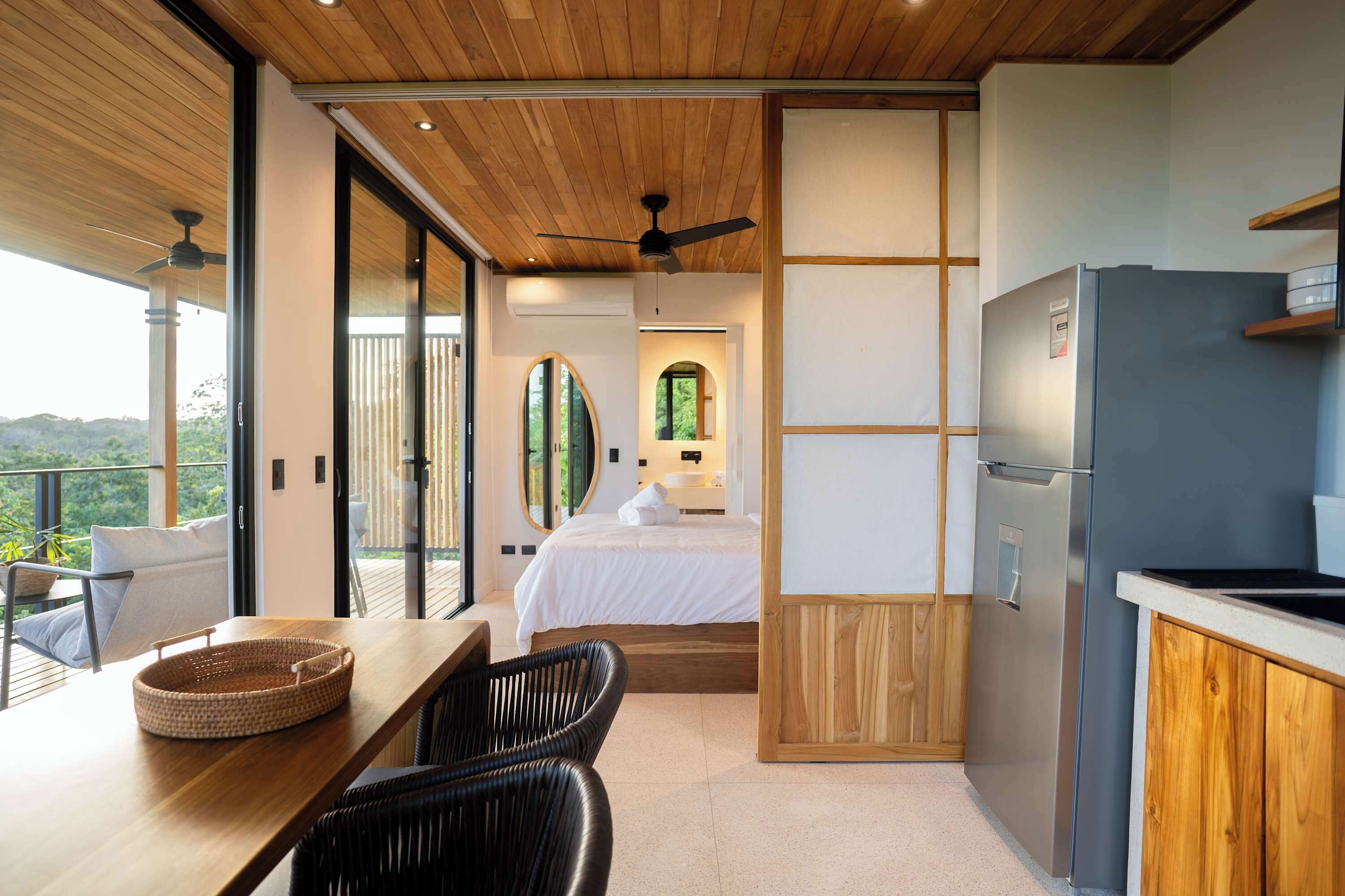A modern bedroom with a large bed, a wooden ceiling fan, and a view of the outdoors.