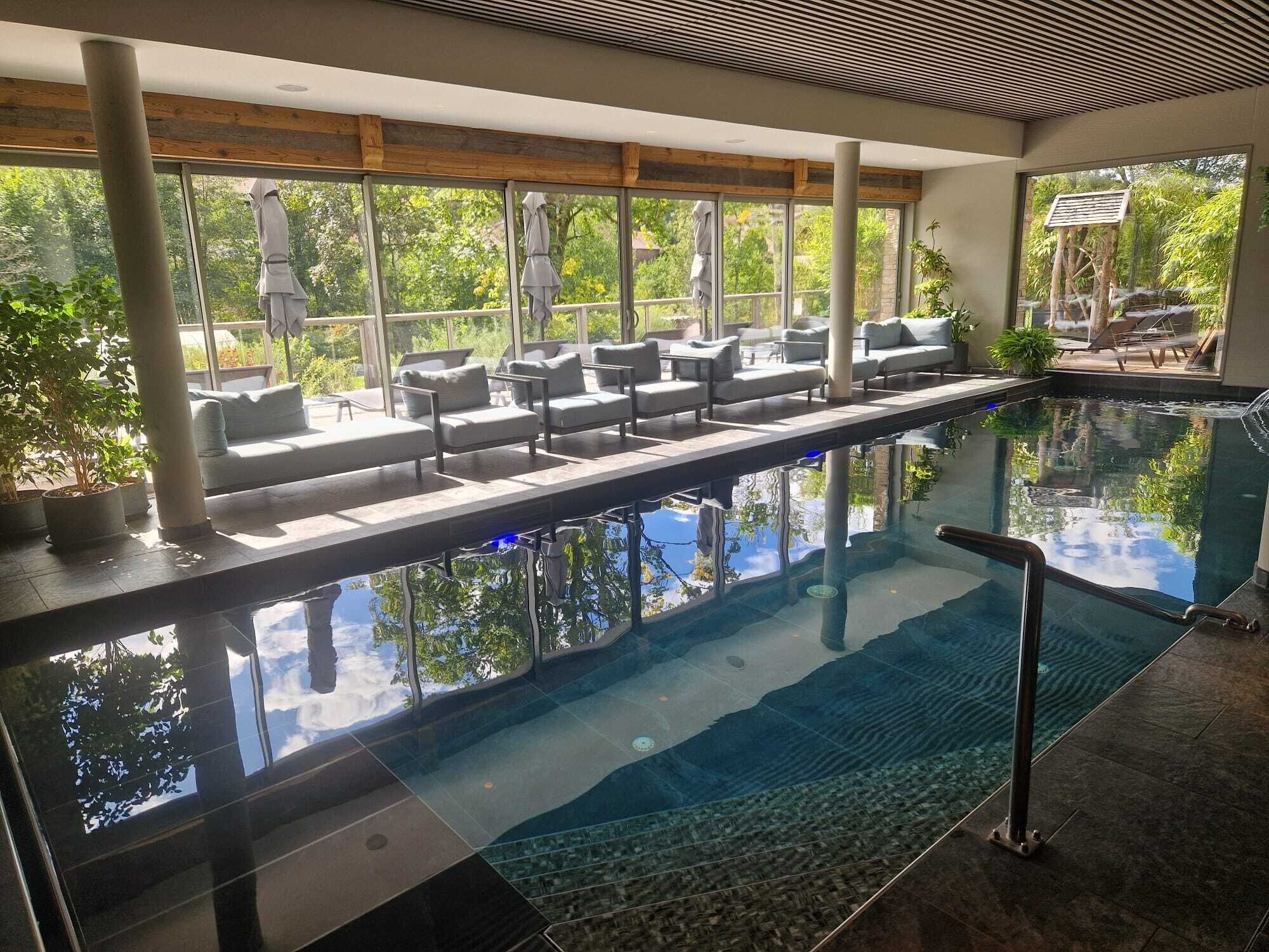 An indoor swimming pool with a view of trees through large windows, a lounge area with sofas, and a stone-tiled floor.