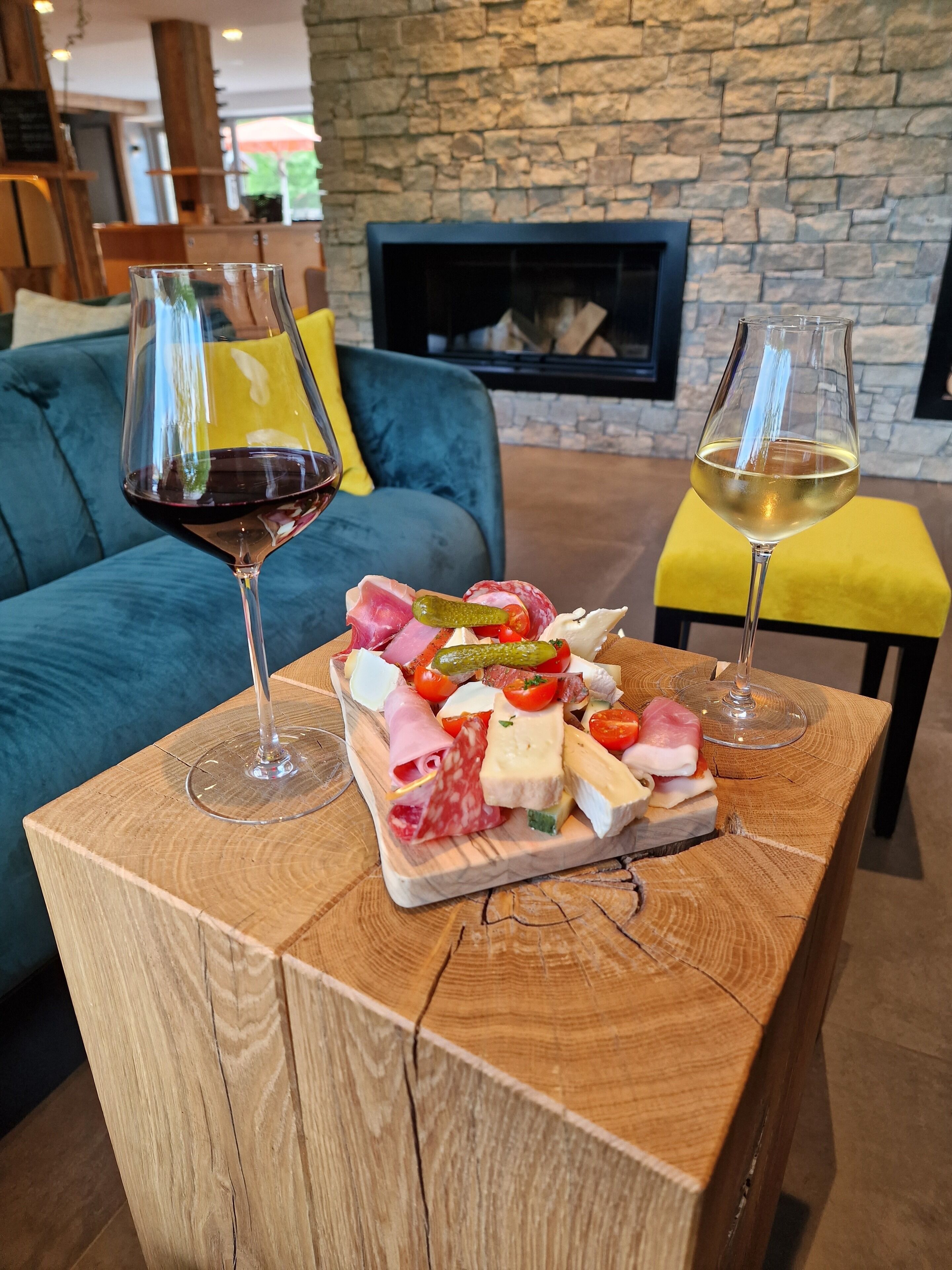 Une table en bois sur laquelle est posée une planche de charcuterie et deux verres à vin, un canapé bleu et, au fond, une cheminée en pierre.