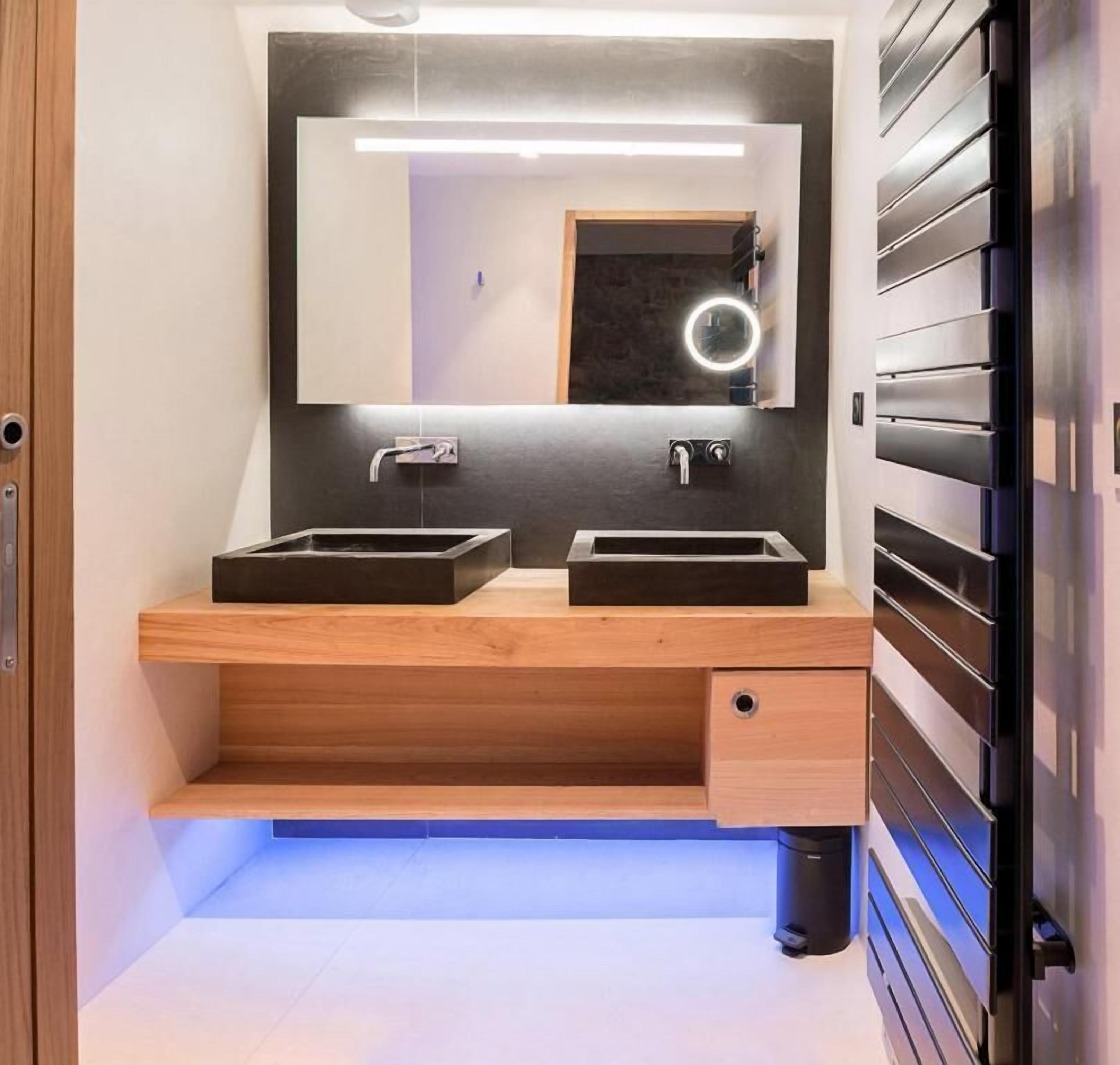 A modern bathroom with two sinks, a mirror with a light, and a wooden vanity with a drawer.