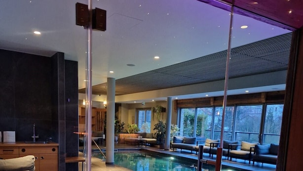 A modern indoor pool area with a wooden deck, seating, and a view through glass walls. - Hôtel Interlaken Lounge Bar & SPA (Xonrupt-Longemer)