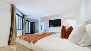 A modern bedroom with a large bed, a flat-screen TV mounted on the wall, and a view of the kitchen through an open door. - Breeze House 2 by Atlantic Holiday (Santa Cruz)