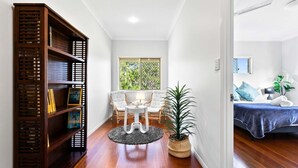 A spacious living room with a wooden bookshelf, a round coffee table, two white cushioned chairs, a large window with a view of greenery, and a bed with a blue bedspread in the adjacent room.
