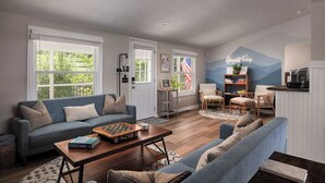 Ein geräumiges Wohnzimmer mit einem blauen Sofa, einem Couchtisch und einem Bücherregal. Eine Tür zeigt eine amerikanische Flagge, eine Wand ist mit einem Bergmotiv gestaltet, und es gibt eine Küchenzeile mit einer Mikrowelle.