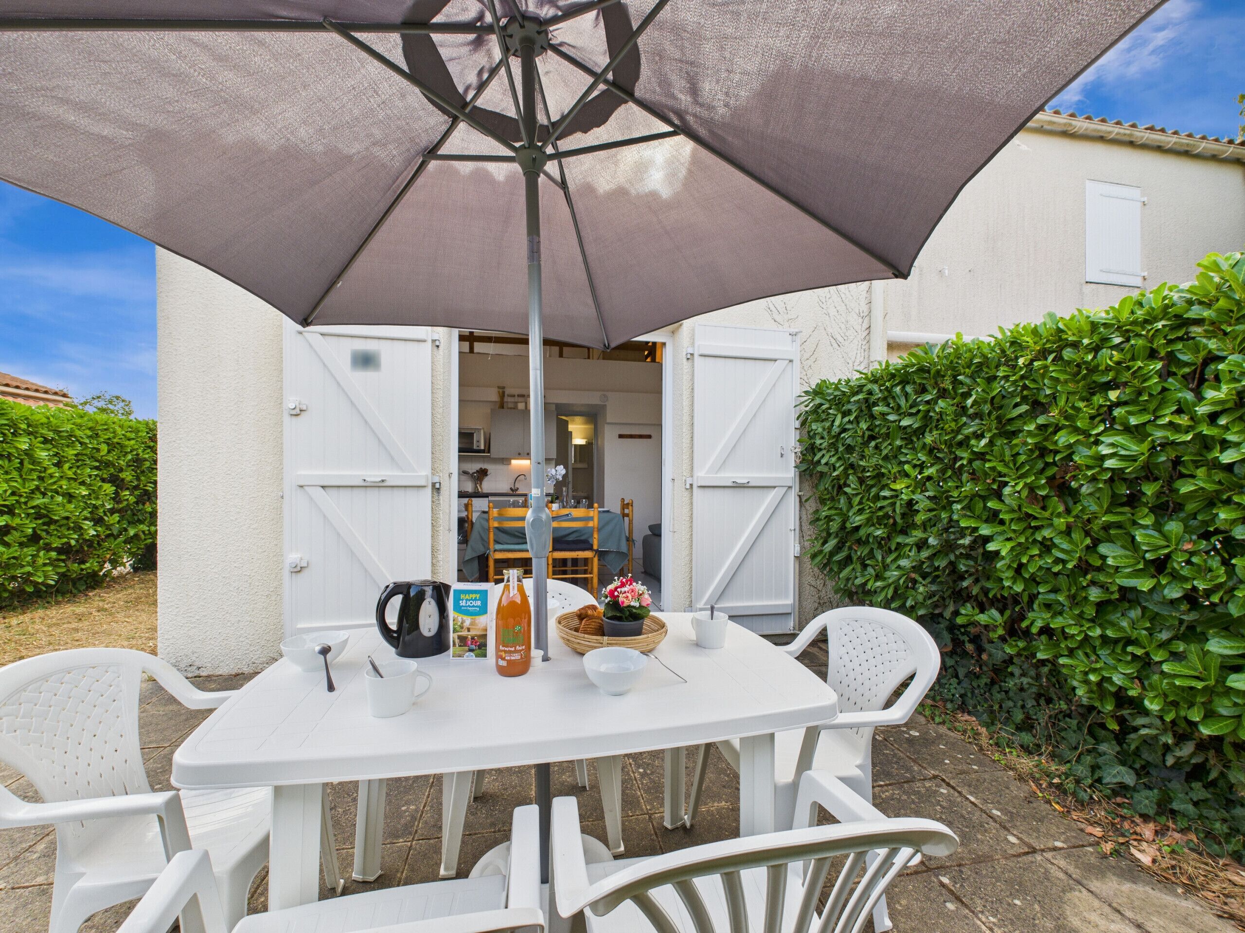 A white table with chairs under a large umbrella, a white door, and a green hedge in the background.