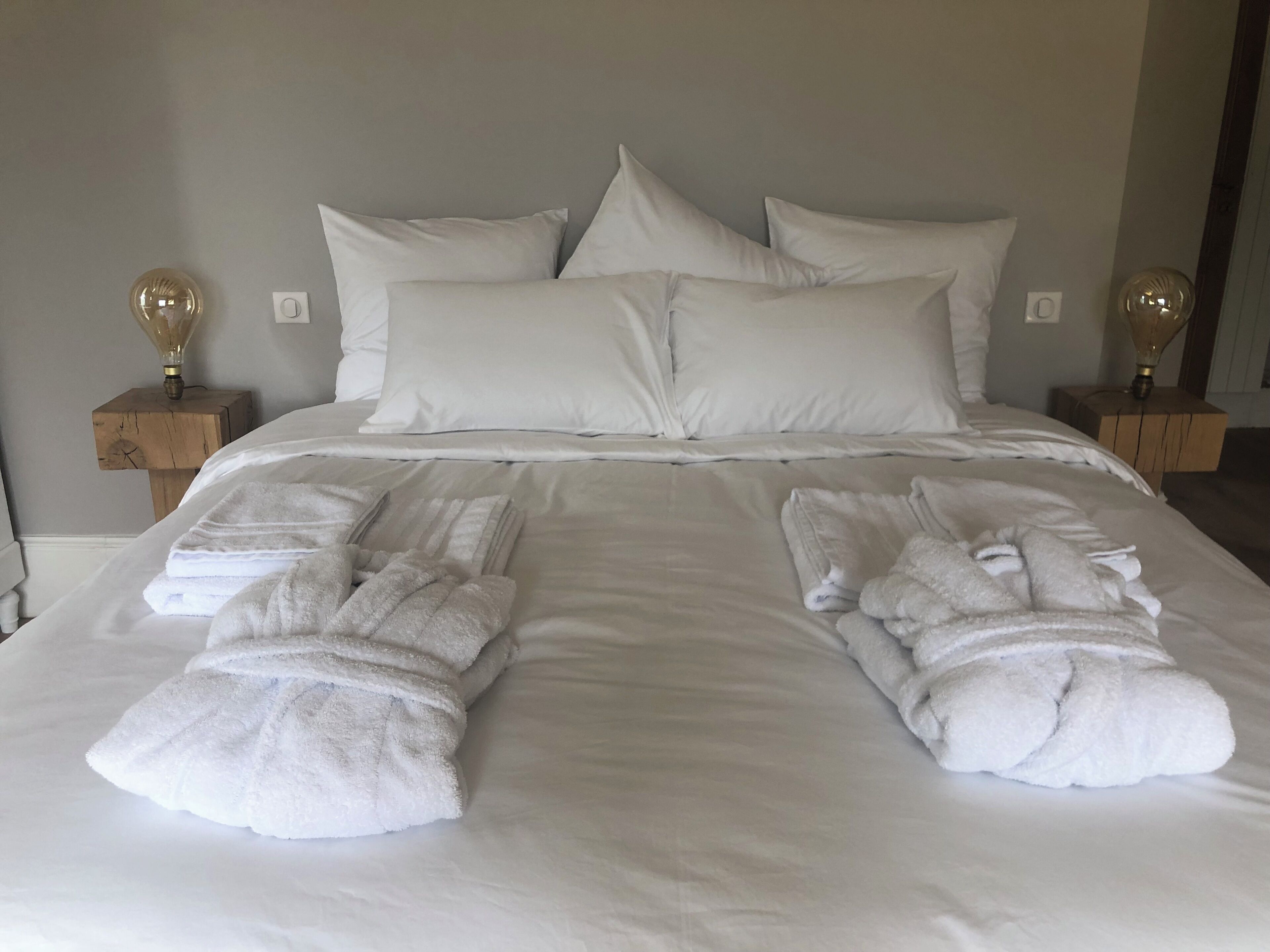 Un lit bien fait, avec du linge de lit blanc et des oreillers, deux tables de chevet avec des lampes, et des serviettes pliées posées sur le lit.