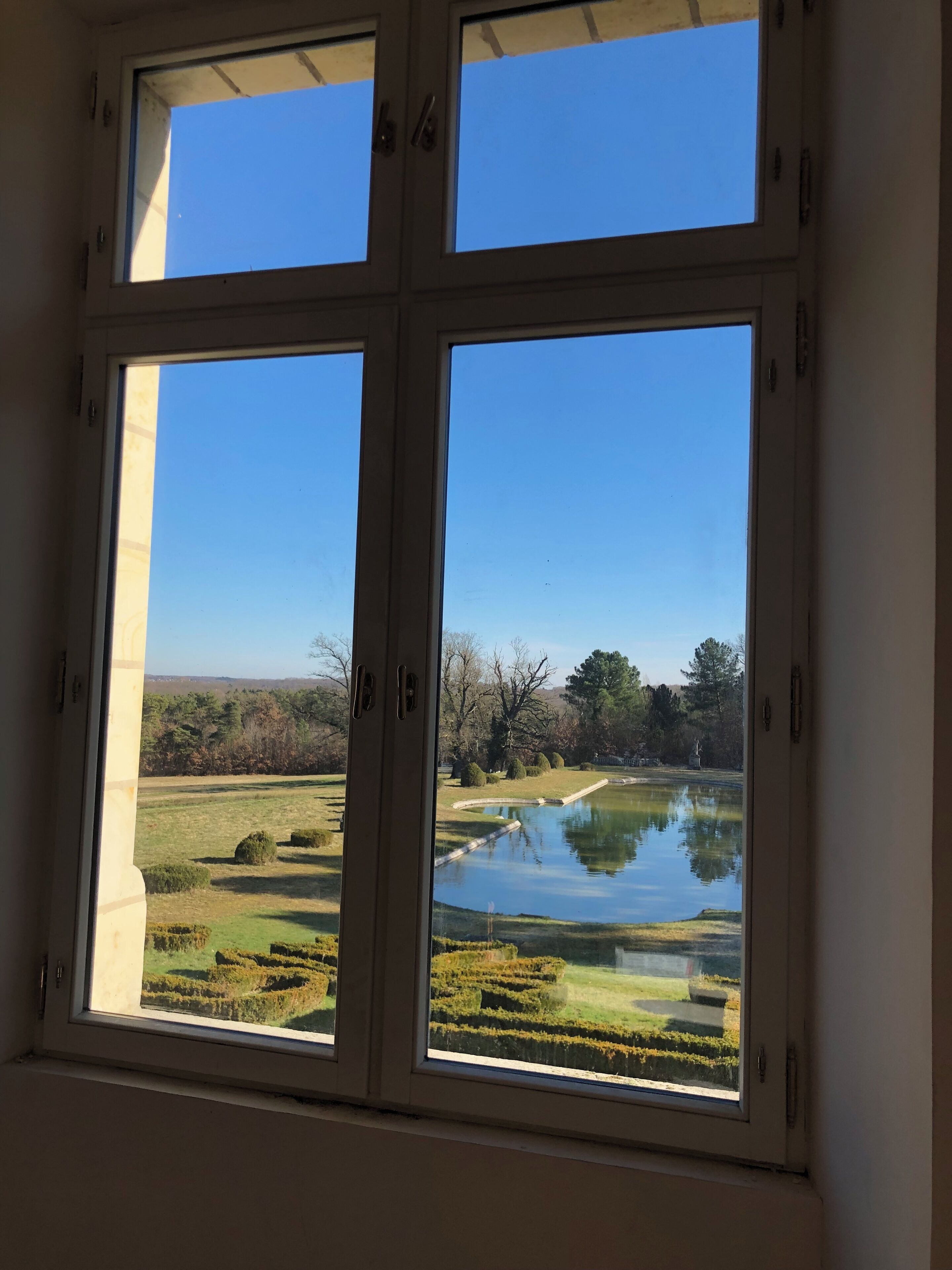 A window with a view of a pond surrounded by neatly trimmed hedges and trees.