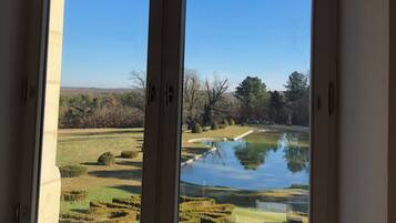 A window with a view of a pond surrounded by neatly trimmed hedges and trees.