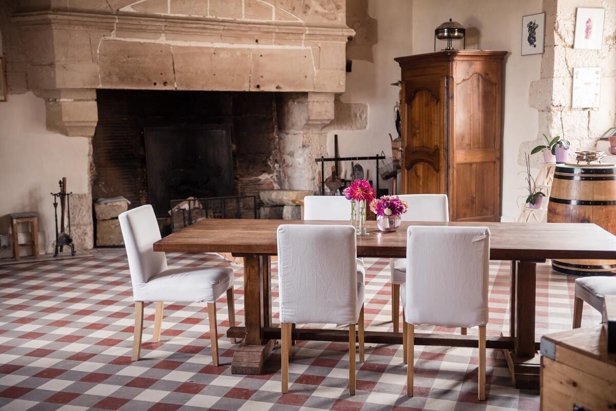 A dining area with a checkered floor, wooden table, white chairs, and a large stone fireplace.