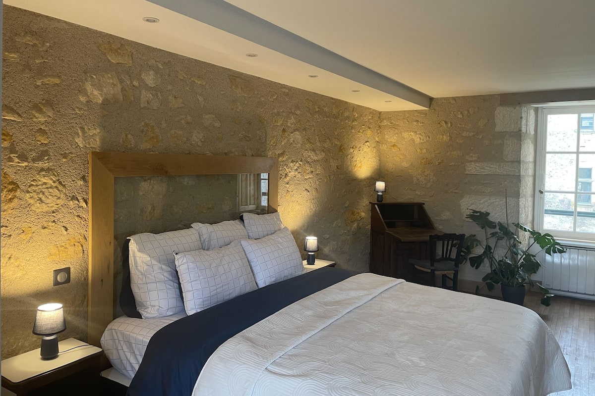 A bedroom with a stone wall, a bed with a dark headboard, bedside lamps, and a window with a view of buildings.