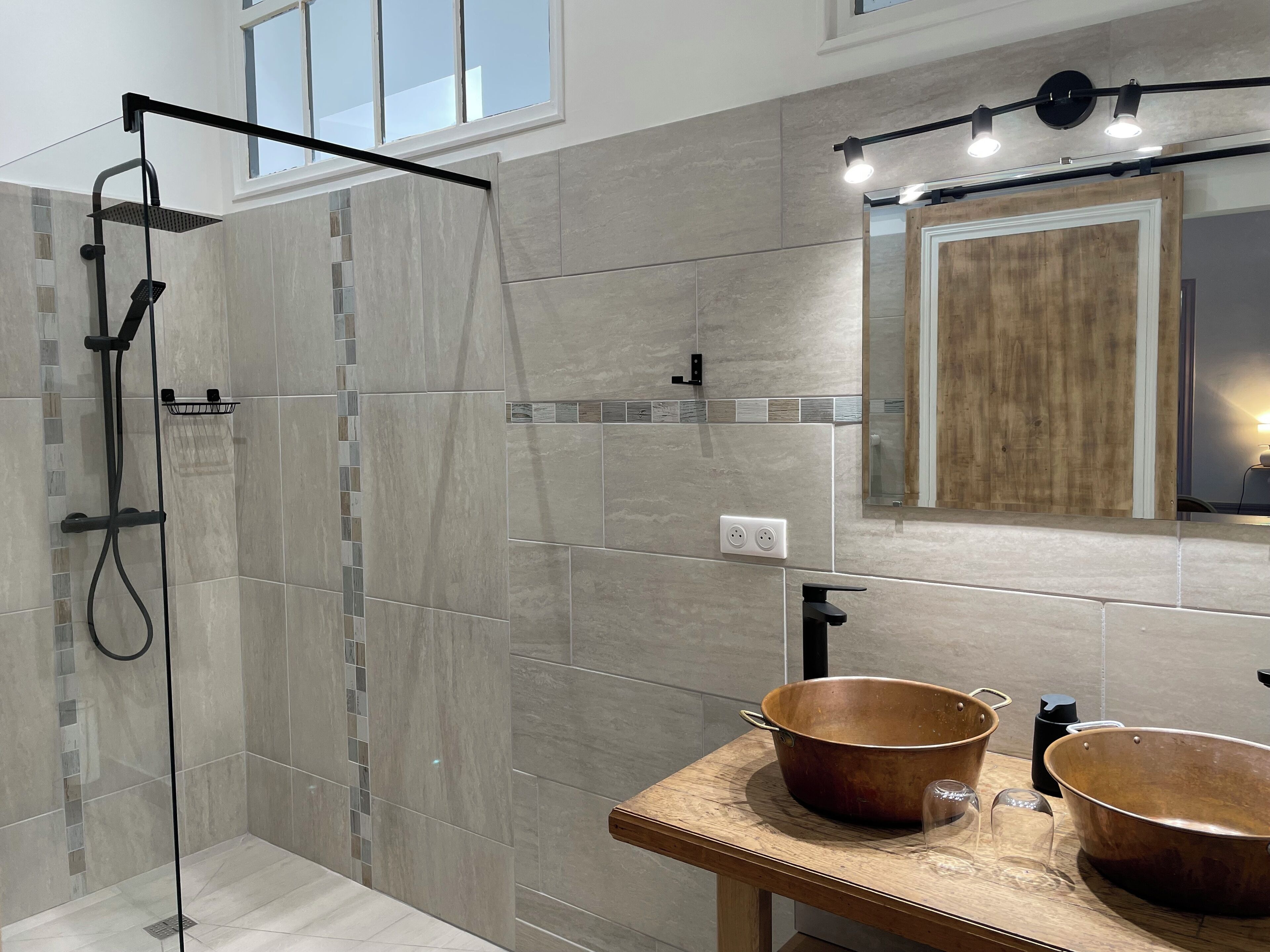 A modern bathroom with a glass-enclosed shower, a wooden vanity with copper sinks, and a mirror with a wooden frame.