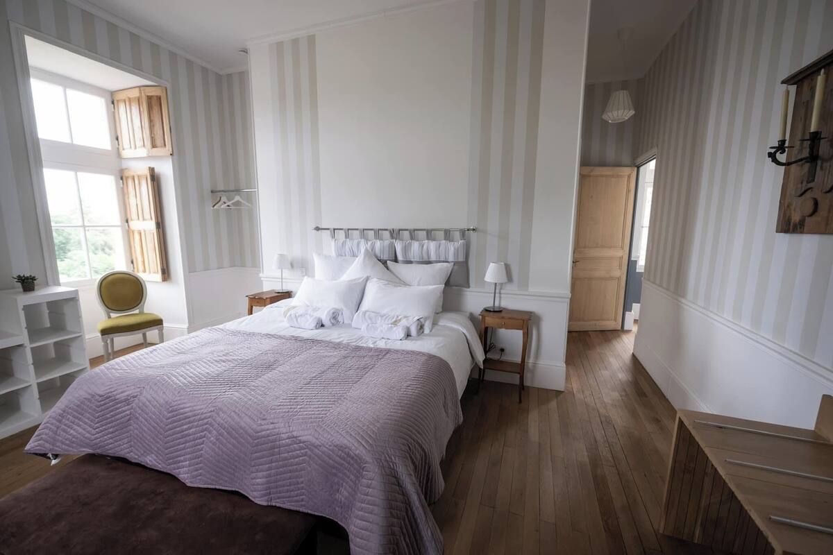 A bedroom with a large bed, wooden flooring, a window with curtains, and a bedside table with a lamp.