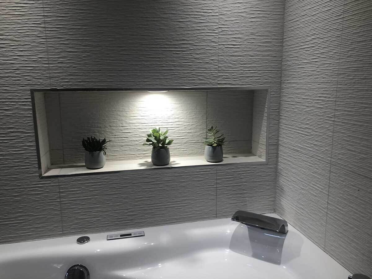 A modern bathroom with a large bathtub, a shelf with three potted plants, and a textured wall.